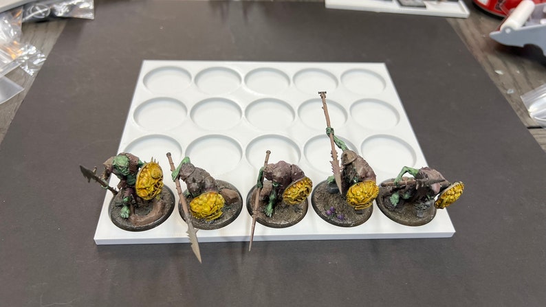 Puede incluir: Cinco figuras en miniatura de color verde y marr&oacute;n con escudos amarillos y lanzas se muestran en una base de pl&aacute;stico blanca con 12 c&iacute;rculos redondos.