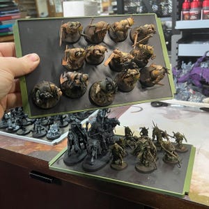 May include: Miniature figurines, painted in shades of brown, tan, and green, are arranged on a black magnetic tray. The figurines, likely for a war game, feature detailed armor and weapons. Another tray with similar figurines is visible below.