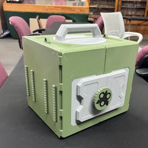 May include: A green and white plastic container with a handle on top and a combination lock design on the front. The container has a boxy shape with a handle and a combination lock design. The color scheme is primarily green and white.