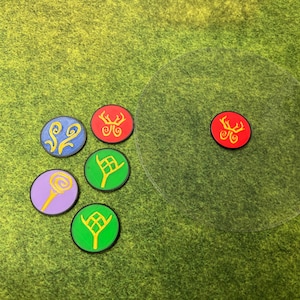 Op de afbeelding: Een verzameling kleurrijke, ronde spel fiches met verschillende symbolen. De fiches hebben een zwarte rand en zijn in tinten blauw, rood, groen en paars. Een doorzichtig, rond spelstuk is ook aanwezig op een groene ondergrond.