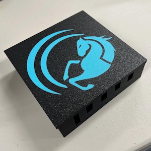 Puede incluir: Caja rectangular negra con superficie texturizada. La parte superior presenta un gráfico azul claro de un caballo encabritado dentro de un diseño curvo. La caja tiene varias aberturas cuadradas en un lado.