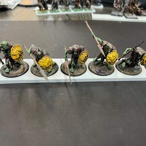 Puede incluir: Cinco figuras en miniatura de orcos pintadas de verde y marr&oacute;n que sostienen lanzas y escudos. Las figuras est&aacute;n sobre una base blanca con c&iacute;rculos negros.