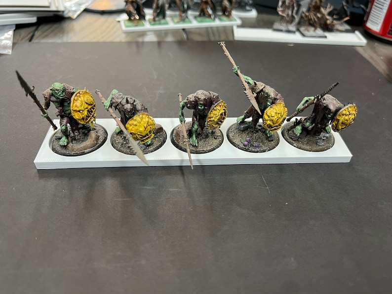 Puede incluir: Cinco figuras en miniatura de orcos pintadas de verde y marr&oacute;n que sostienen lanzas y escudos. Las figuras est&aacute;n sobre una base blanca con c&iacute;rculos negros.