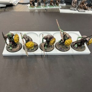 Puede incluir: Cinco figuras en miniatura de orcos pintadas de verde y marr&oacute;n que sostienen lanzas y escudos. Las figuras est&aacute;n de pie sobre una base de pl&aacute;stico blanca con puntos negros.