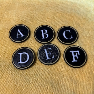 Op de afbeelding: Zes ronde zwarte en gouden tegels met witte letters die het woord "ABCDEF" spellen.