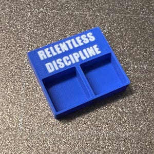 Puede incluir: Un organizador rectangular azul con dos compartimentos. Las palabras "RELENTLESS DISCIPLINE" están impresas en blanco en la parte superior. El organizador está hecho de un material mate y sólido.