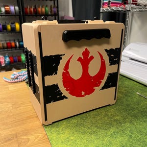 May include: A tan box with a black handle and accents. The box features a red and black graphic of a symbol. The box is sitting on a wooden surface.