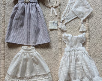 Op de afbeelding: Een set poppenkleding inclusief een gestreepte jurk, een witte jurk met een schort, een witte rok, een witte kanten sjaal en een witte gehaakte ketting.