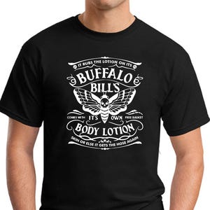 May include: Black t-shirt featuring a white graphic design. The design includes the text "IT RUBS THE LOTION ON ITS BUFFALO BILL'S BODY LOTION" with a moth illustration. The shirt has short sleeves and a crew neck.