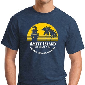 May include: A navy blue t-shirt featuring a yellow graphic of a lighthouse, palm trees, and a sunset. The text "Amity Island, New England, USA" is printed below the graphic. The text "Clear Skies, Gentle Surf, Warm Waters" is printed in a circle around the graphic.