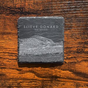 May include: A square, dark grey slate coaster featuring the text "SLIEVE DONARD 850 METRES" and a mountain illustration. The coaster rests on a wooden surface with a warm, brown colour.