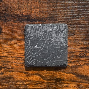 Puede incluir: Un posavasos de pizarra negra con un diseño de mapa topográfico blanco y la palabra "HAYSTACKS" impresa en él.