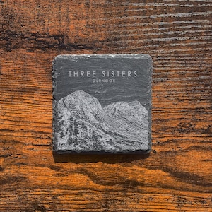 May include: A black slate coaster featuring a white image of the Three Sisters mountains in Glencoe, Scotland. The text "THREE SISTERS GLENCOE" is printed on the coaster.