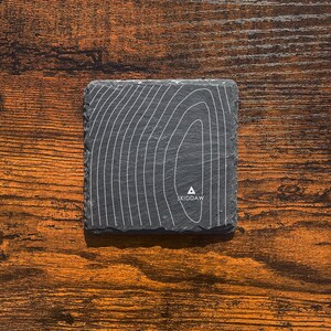 May include: A square, dark grey slate coaster featuring a white topographic map design. The word "SKIDDAW" is printed in white at the bottom. The coaster is placed on a wooden surface.