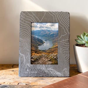 May include: A grey slate photo frame featuring a white topographic map design. The frame displays a landscape photo of a lake and mountains. The frame sits on a wooden surface with a succulent plant and a leather-bound notebook.
