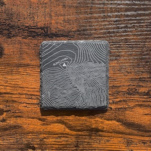 May include: A black slate coaster featuring a topographic map of the mountain "Blencathra". The map is printed in white and shows the contours of the mountain.