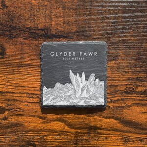 May include: A square, dark grey slate coaster featuring the text "GLYDER FAWR" and "1001 METRES" in white. A detailed white illustration of a mountain range is below the text. The coaster is on a wooden surface.