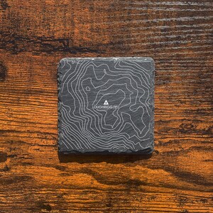 May include: A black slate coaster featuring a topographic map design of Loughrigg Fell. The coaster has white lines depicting the contours of the mountain.