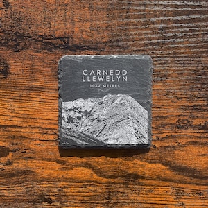 May include: A black slate coaster with a white image of a mountain peak and the text "Carnedd Llewelyn 1062 Metres".
