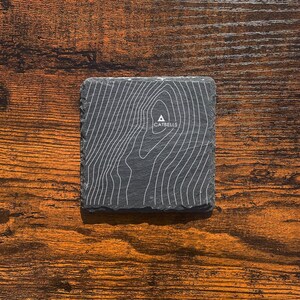 May include: A black slate coaster featuring a topographic map design with the word "Catbells" printed in white.