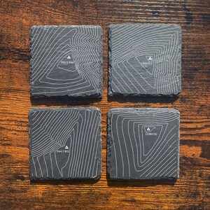 以下が含まれることがあります： 黒曜石製のコースター4枚セット。白い地形図で山が描かれています。コースターごとに異なる山が描かれており、それぞれに名前が書かれています。Pen y Fan、Cribyn、Fan y Big、Corn Duです。