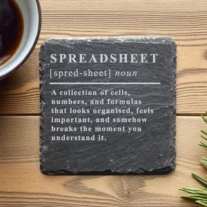 Könnte beinhalten: Ein quadratischer, dunkelgrauer Schieferuntersetzer mit dem Wort "SPREADSHEET" in Weiß und seiner Definition. Der Untersetzer liegt auf einer Holzoberfläche, mit einer Tasse Kaffee und Kiefernnadeln im Hintergrund.