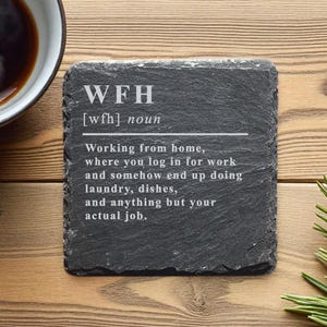 Könnte beinhalten: Ein schwarzer Schieferuntersetzer mit dem Text "WFH [wfh] noun" und einer Definition von Homeoffice. Der Untersetzer liegt auf einer Holzoberfläche neben einer Tasse Kaffee und einem Zweig Grün.