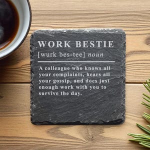 Könnte beinhalten: Ein quadratischer, dunkelgrauer Schieferuntersetzer mit dem weißen Text "WORK BESTIE". Die Definition von "work bestie" ist darunter abgedruckt. Links befindet sich eine Tasse Kaffee, rechts Kiefernnadeln.