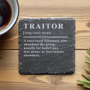 Könnte beinhalten: Ein quadratischer, schwarzer Schieferuntersetzer mit der Definition von "TRAITOR" in weißer Schrift. Die Definition enthält die Aussprache und eine humorvolle Erklärung. Eine Tasse Kaffee ist links zu sehen.