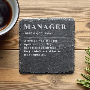 Könnte beinhalten: Ein quadratischer, dunkelgrauer Schieferuntersetzer mit dem Wort "MANAGER" in Weiß und einer Definition. Der Untersetzer liegt auf einer Holzoberfläche, neben einer Tasse Kaffee und einem Kiefernzweig.