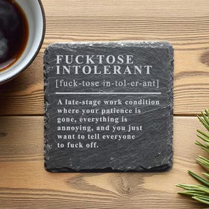 Könnte beinhalten: Ein schwarzer Schieferuntersetzer mit dem Text "FUCKTOSE INTOLERANT" und einer Definition. Links steht eine Tasse Kaffee, rechts ein Kiefernzweig. Der Untersetzer liegt auf einer Holzoberfläche.