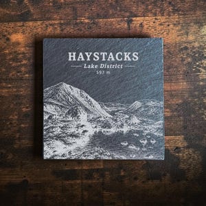 Può includere: Un sottobicchiere quadrato in ardesia grigio scuro con un'illustrazione bianca incisa della montagna Haystacks nel Lake District. Il testo riporta HAYSTACKS, Lake District e 597 m. La superficie presenta una finitura naturale, opaca e ruvida.