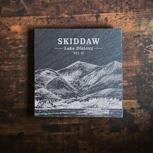 May include: A square dark grey slate coaster featuring a white etched illustration of the Skiddaw mountain range in the Lake District. The text reads SKIDDAW, Lake District, and 931 m, depicting a landscape with a boat on the water.