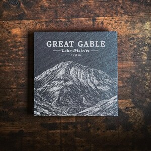 May include: A square slate coaster featuring a white engraved illustration of the Great Gable mountain in the Lake District. The text reads GREAT GABLE, Lake District, and 899 m. The coaster has a dark grey, textured surface with natural stone edges.