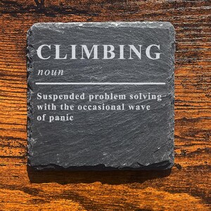 May include: A square, dark grey slate coaster with the word "CLIMBING" in white capital letters. Below, the definition reads: "Suspended problem solving with the occasional wave of panic."