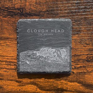 May include: A black slate coaster with the words "Clough Head 724 Metres" printed in white. The coaster features a line drawing of a mountain.