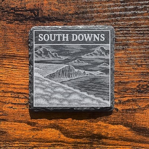 Può includere: Sottobicchiere quadrato in ardesia con una scena di paesaggio incisa e le parole "SOUTH DOWNS" in un bordo rettangolare. Il sottobicchiere è posto su una superficie di legno scuro.
