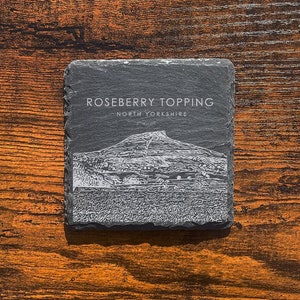May include: A black slate coaster with a white image of Roseberry Topping, a hill in North Yorkshire, England. The text "Roseberry Topping" and "North Yorkshire" is printed in white on the coaster.