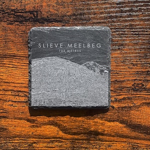 May include: A square, dark grey slate coaster featuring the text "SLIEVE MEELBEG" and "702 METRES" in white. A grayscale image of a mountain is below the text. The coaster rests on a wooden surface.