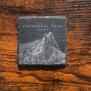 May include: Square, dark grey slate coaster featuring the text "CATHEDRAL PEAK YOSEMITE" and a white illustration of a mountain. The coaster is on a wooden surface with a brown hue.