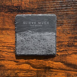 Może przedstawiać: Kwadratowa, szara podstawka z łupków z białym napisem "SLIEVE MUCK" i "670 METRES". Podstawka ma wzór górski. Podstawka znajduje się na drewnianej powierzchni w ciepłym, brązowym kolorze.