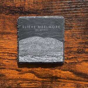May include: A square, dark gray slate coaster with the text "SLIEVE MEELMORE" and "680 METRES" in white. The coaster features a grayscale image of a mountain range. The coaster is on a wooden surface.