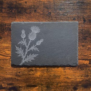May include: Rectangular black slate serving board with a white etched thistle design. The board is set against a dark wood grain background. The thistle design is on the left side of the board.