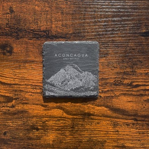 May include: A black slate coaster featuring a white outline of Aconcagua, the highest mountain in the Andes, with the text "Aconcagua 6961 Metres".