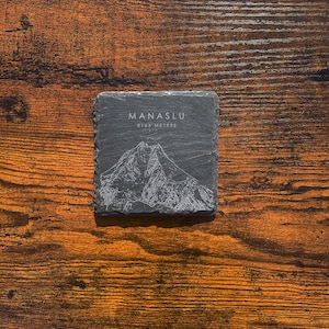 May include: Square, dark gray slate coaster with the name "MANASLU" and "8163 METRES" etched in white. A detailed mountain illustration is also etched on the coaster. The coaster sits on a wooden surface.