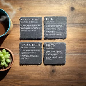 May include: Four slate coasters with white text describing the Lake District, Fell, Wainwright, and Beck. The coasters are arranged on a wooden surface, with a cup of coffee and a small succulent plant visible.