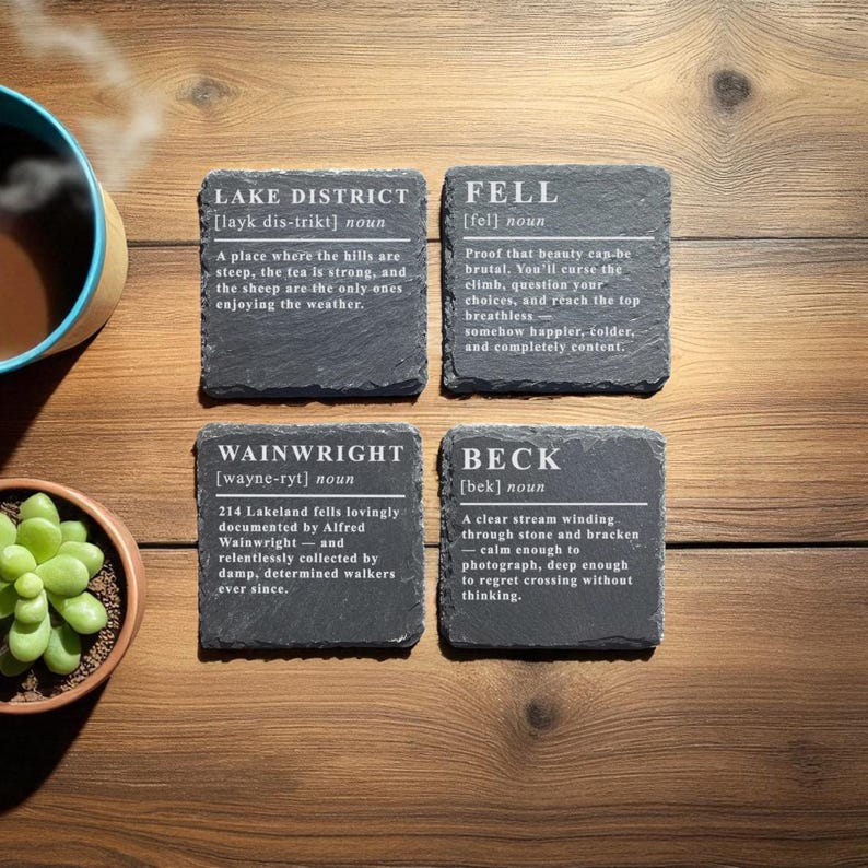 May include: Four slate coasters with white text describing the Lake District, Fell, Wainwright, and Beck. The coasters are arranged on a wooden surface, with a cup of coffee and a small succulent plant visible.