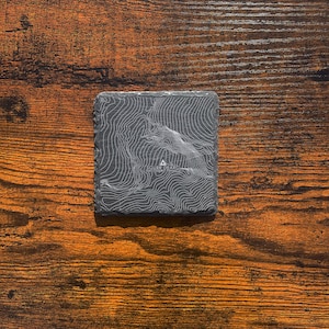 May include: A black slate coaster featuring a topographic map of Scurr Dearg, a mountain in Ireland. The coaster is on a wooden surface.