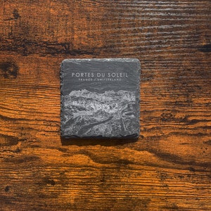 May include: A black slate coaster with the text "Portes du Soleil" and "France/Switzerland" in white lettering. The coaster features a white etched image of a mountain range.