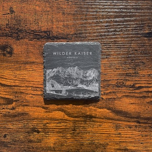May include: A black slate coaster with a white etching of a mountain range and the text "Wilder Kaiser Austria".